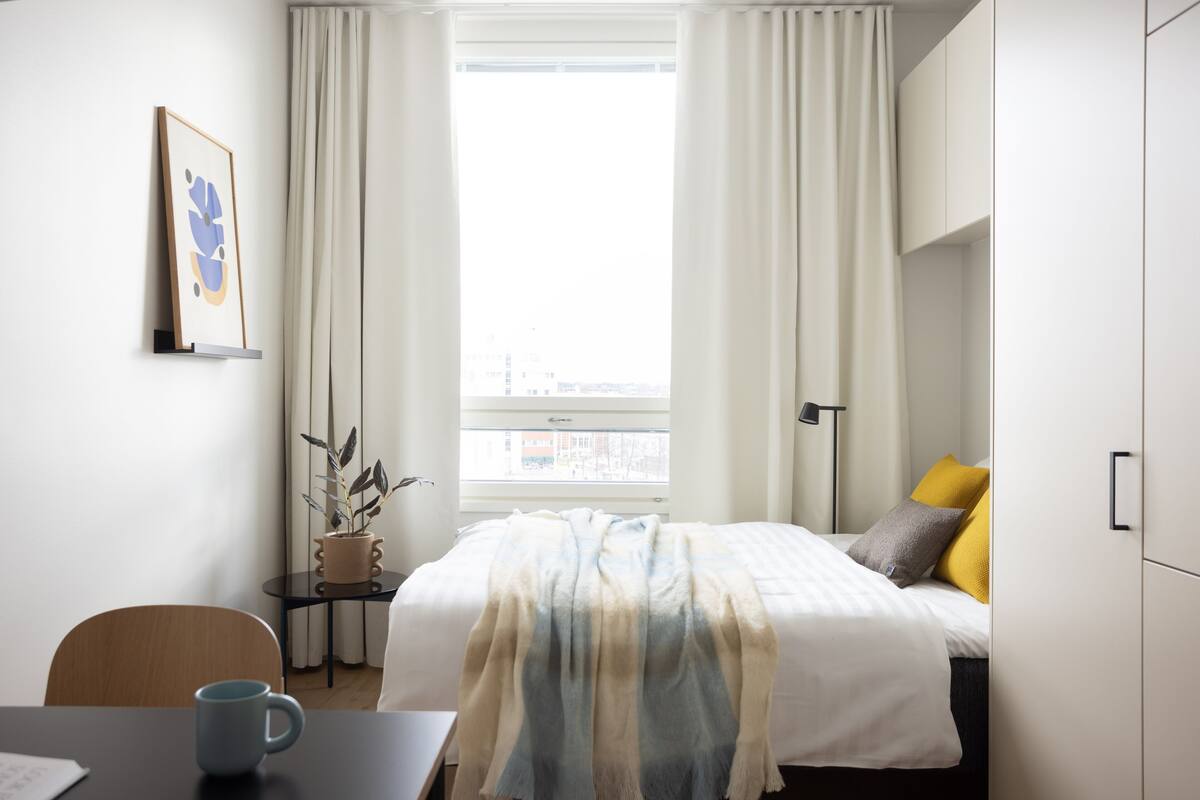 Quarto de hotel com cama, escrivaninha, cadeira, janela com cortinas, planta e quadro na parede.