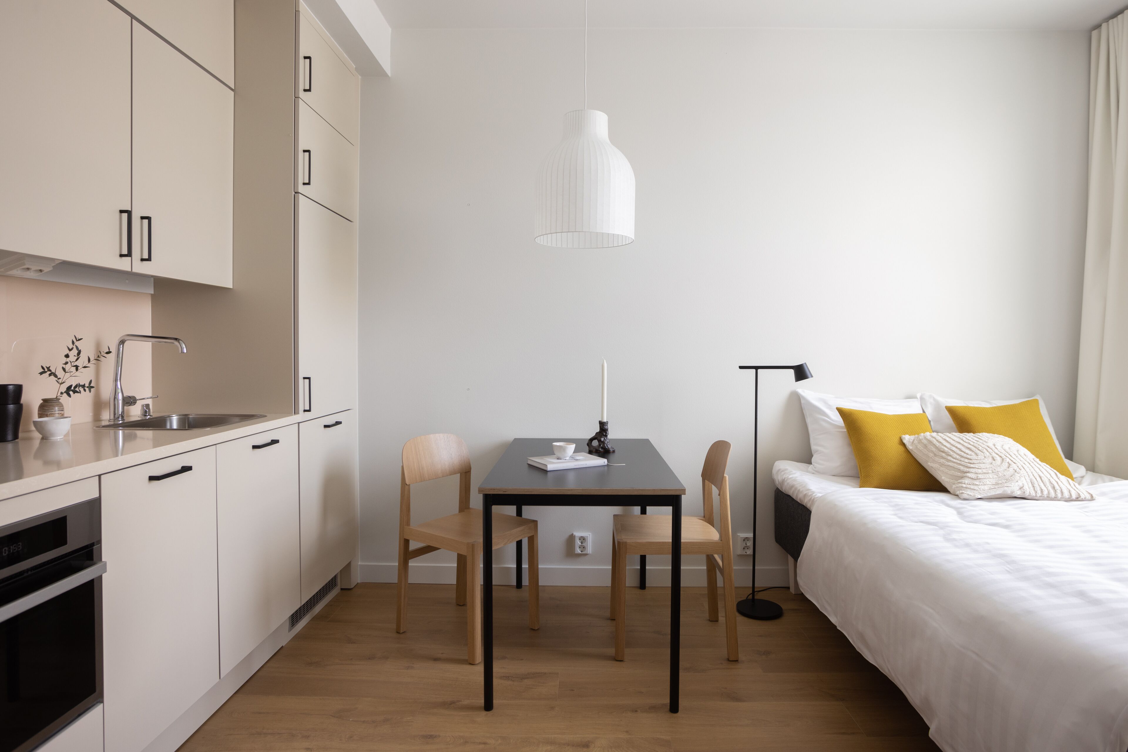 Una habitación de hotel moderna con kitchenette, un comedor con dos sillas, una cama con almohadas blancas y amarillas, y una lámpara colgante.