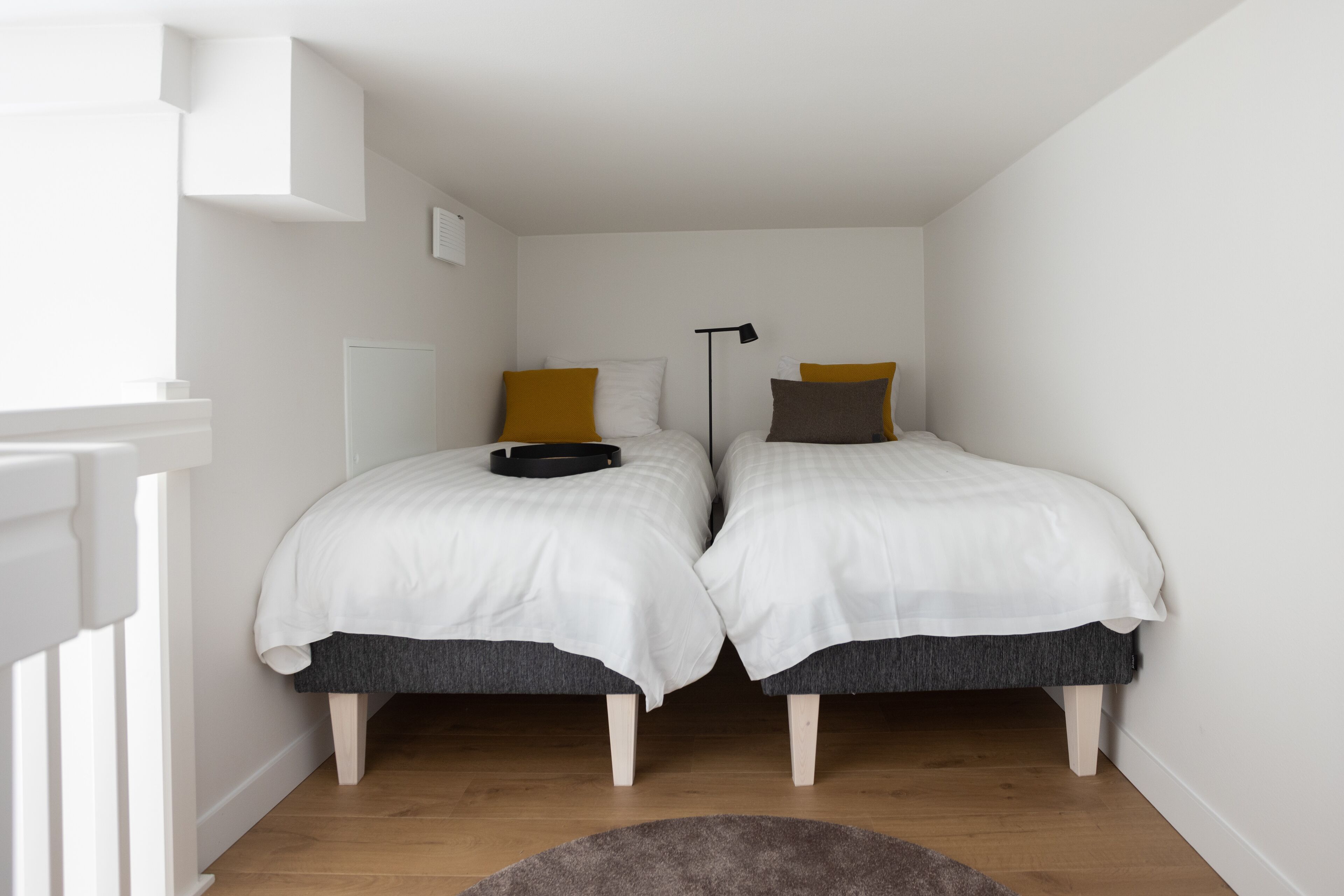 A small, well-lit bedroom with two single beds, a bedside table, a floor lamp, and a wooden floor.