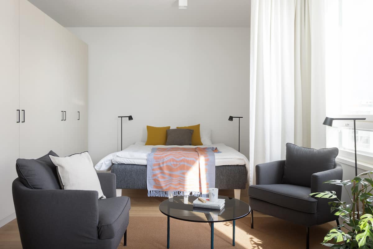 Quarto de hotel moderno com uma cama, duas poltronas, uma mesa pequena e uma janela ampla com cortinas.