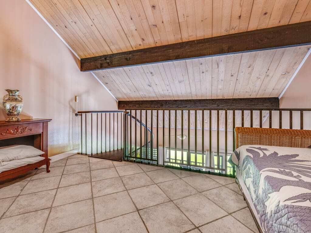 Quarto sótão com pé-direito alto, chão de madeira, cama, mesa de cabeceira e escada que dá para área externa.