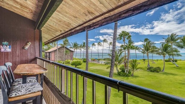 A balcony with a wooden table and chairs, overlooking a lawn with palm trees and a view of the ocean.