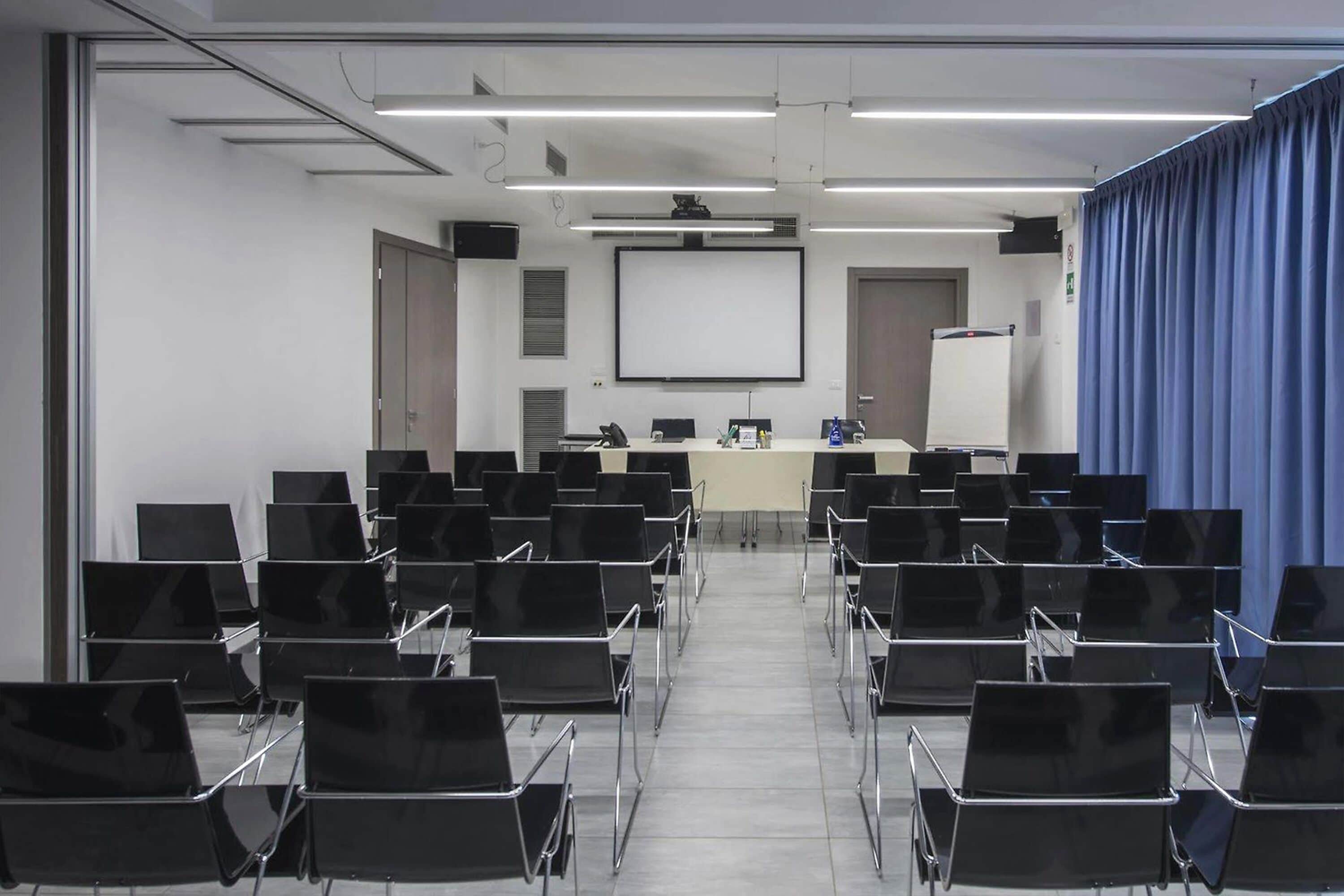 A conference room with rows of black chairs, a projector screen, a whiteboard, and a blue curtain.