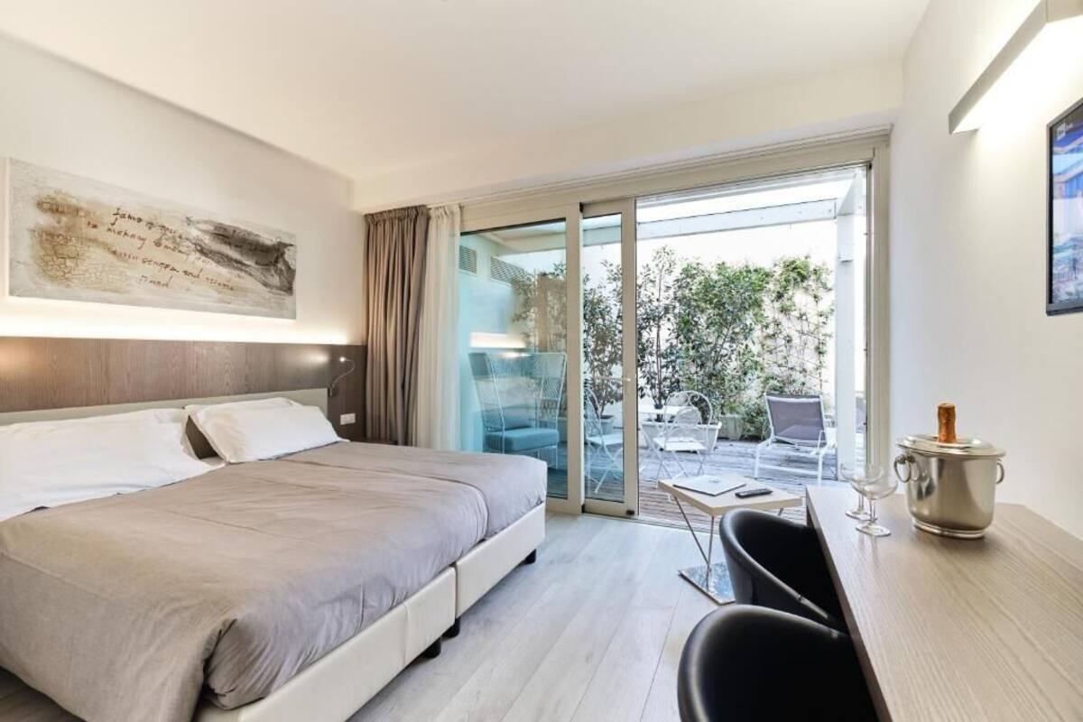 A modern hotel room with a bed, a desk with a chair, a large window, and a sliding glass door leading to an outdoor area.