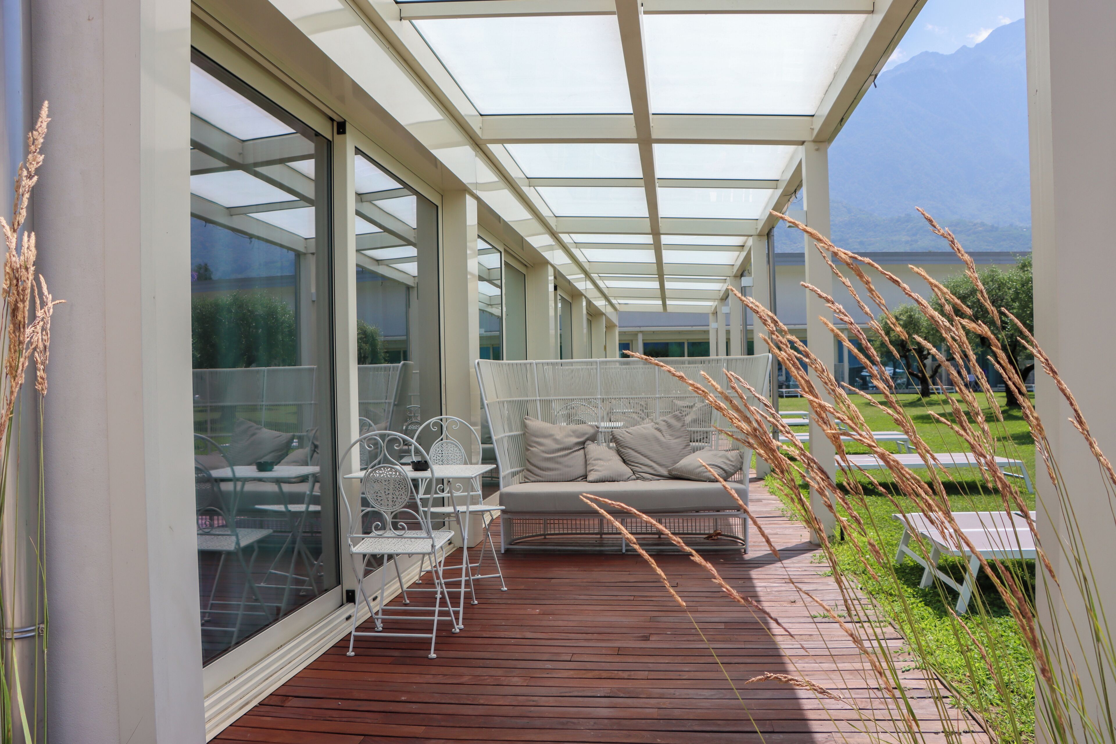 A covered outdoor seating area with a wooden deck, white metal chairs, and a cushioned bench.