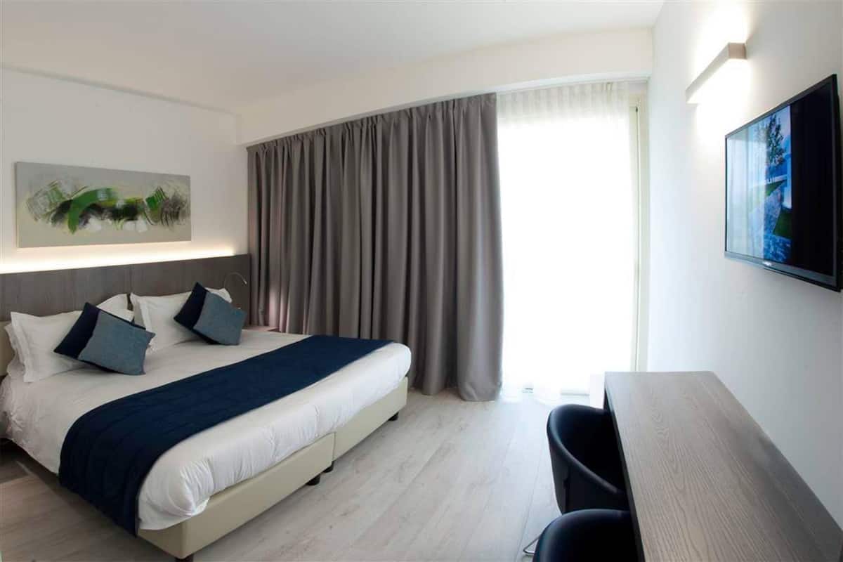 A modern hotel room with a bed, a desk, and a large window with curtains.