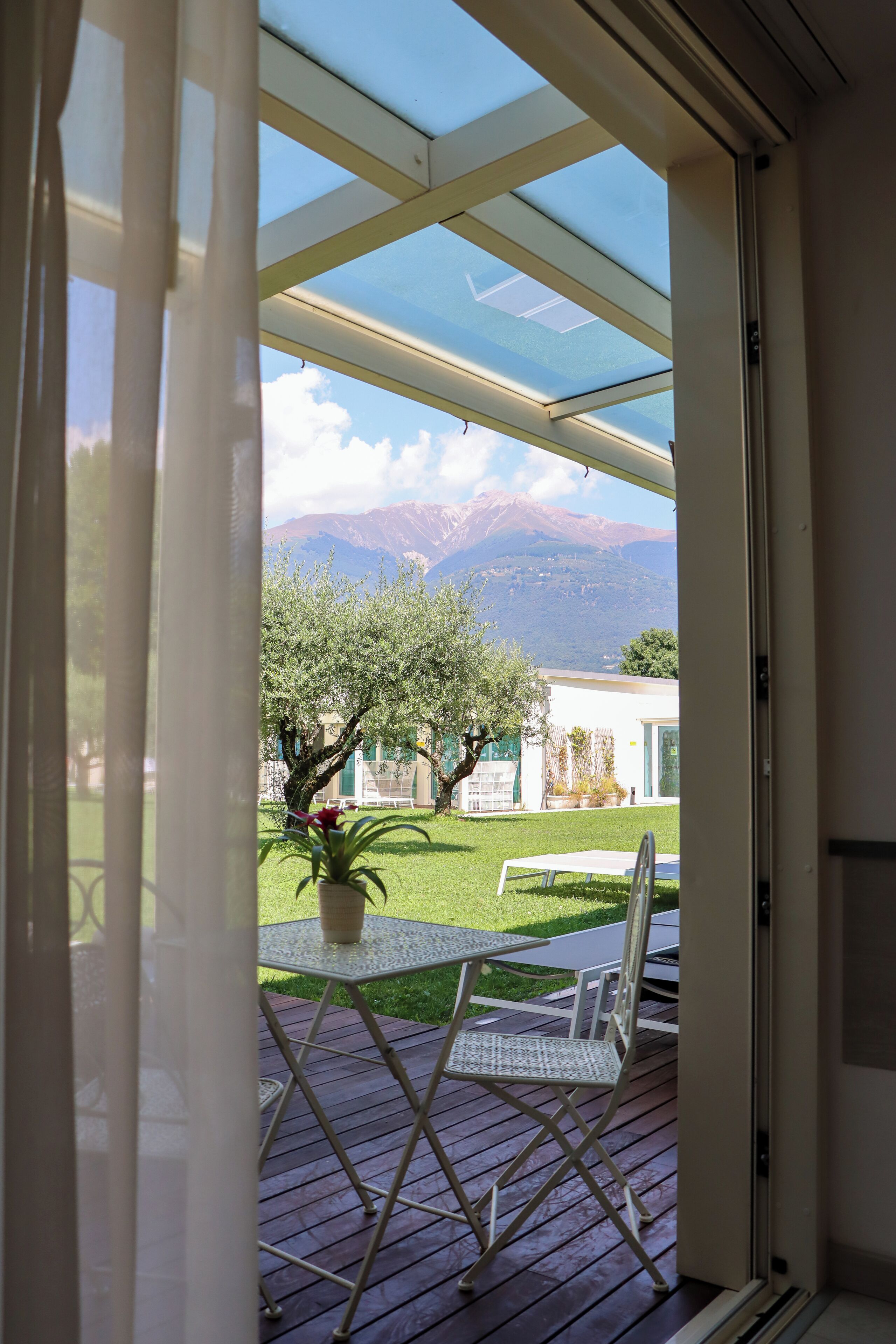 Vista a través de un ventanal a un patio con una mesa, sillas, una planta en maceta y una montaña al fondo.