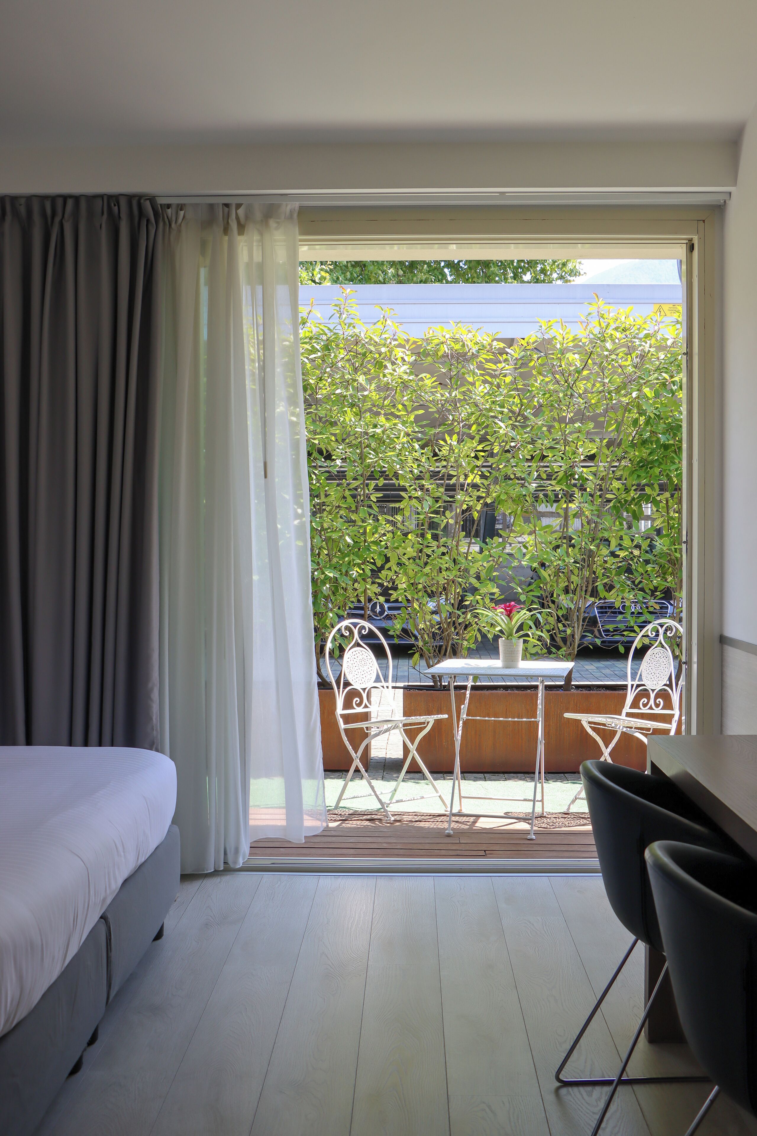 A bedroom with a bed, a table with chairs, and a view of a balcony with plants and a car.