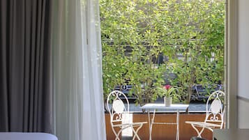 A bedroom with a bed, a table with chairs, and a view of a balcony with plants and a car.