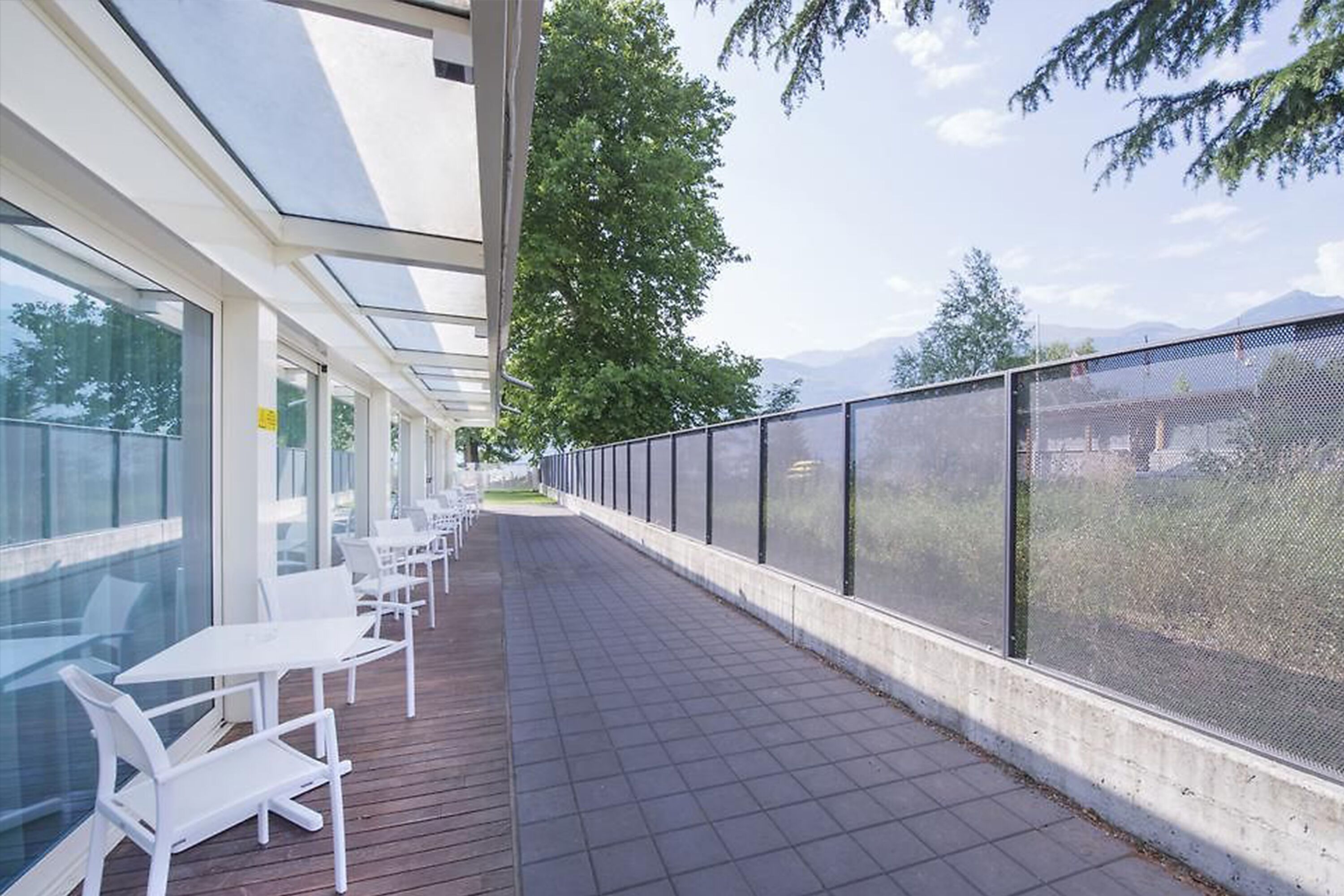 A covered outdoor seating area with white chairs and tables, a glass wall, and a view of trees and mountains.