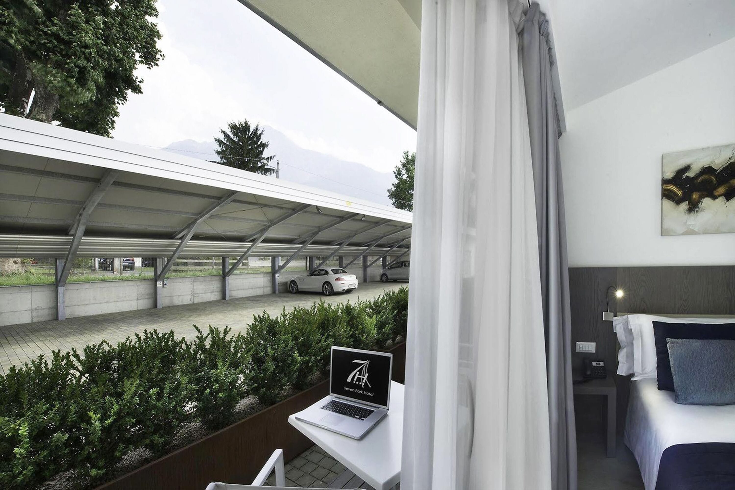 Ein modernes Hotelzimmer mit einem Laptop auf dem Tisch, einem Bett mit Kissen, einem Bild an der Wand und Blick auf einen Parkplatz mit Autos und Bergen im Hintergrund.