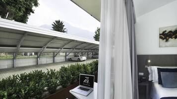 Ein modernes Hotelzimmer mit einem Laptop auf dem Tisch, einem Bett mit Kissen, einem Bild an der Wand und Blick auf einen Parkplatz mit Autos und Bergen im Hintergrund.