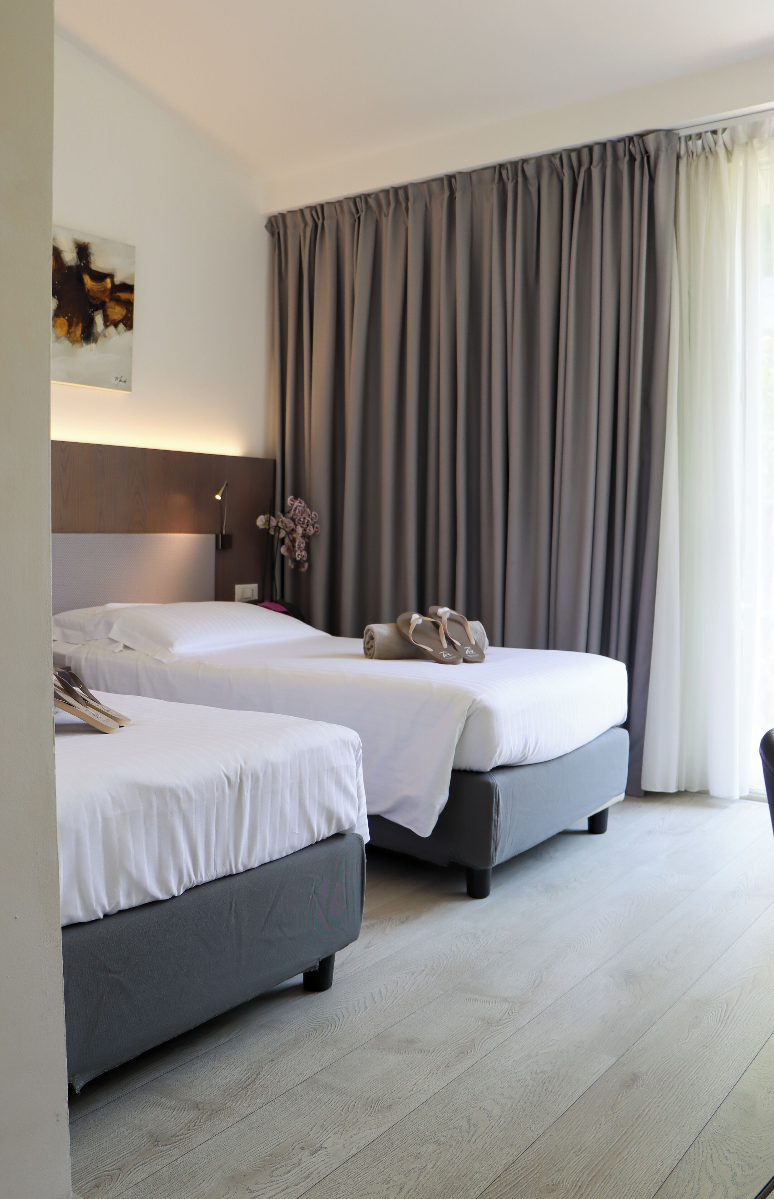 Ein Hotelzimmer mit zwei Betten, einem großen Fenster mit Vorhängen, einer Wandlampe und einem Gemälde.