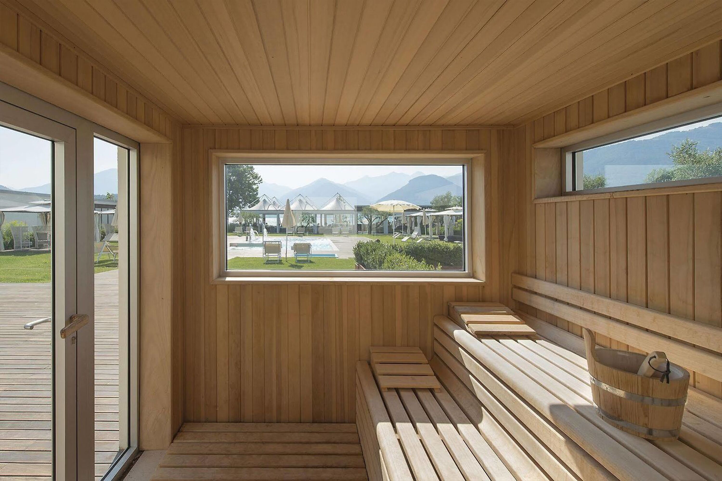 A sauna with wooden benches and a large window offering a view of a pool and mountains.