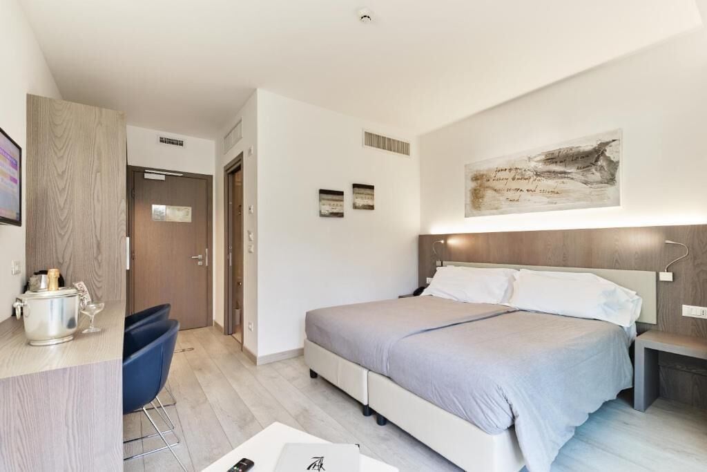 A modern hotel room with a bed, bedside table, and a desk with a chair. There is a painting on the wall and a small table with a lamp.