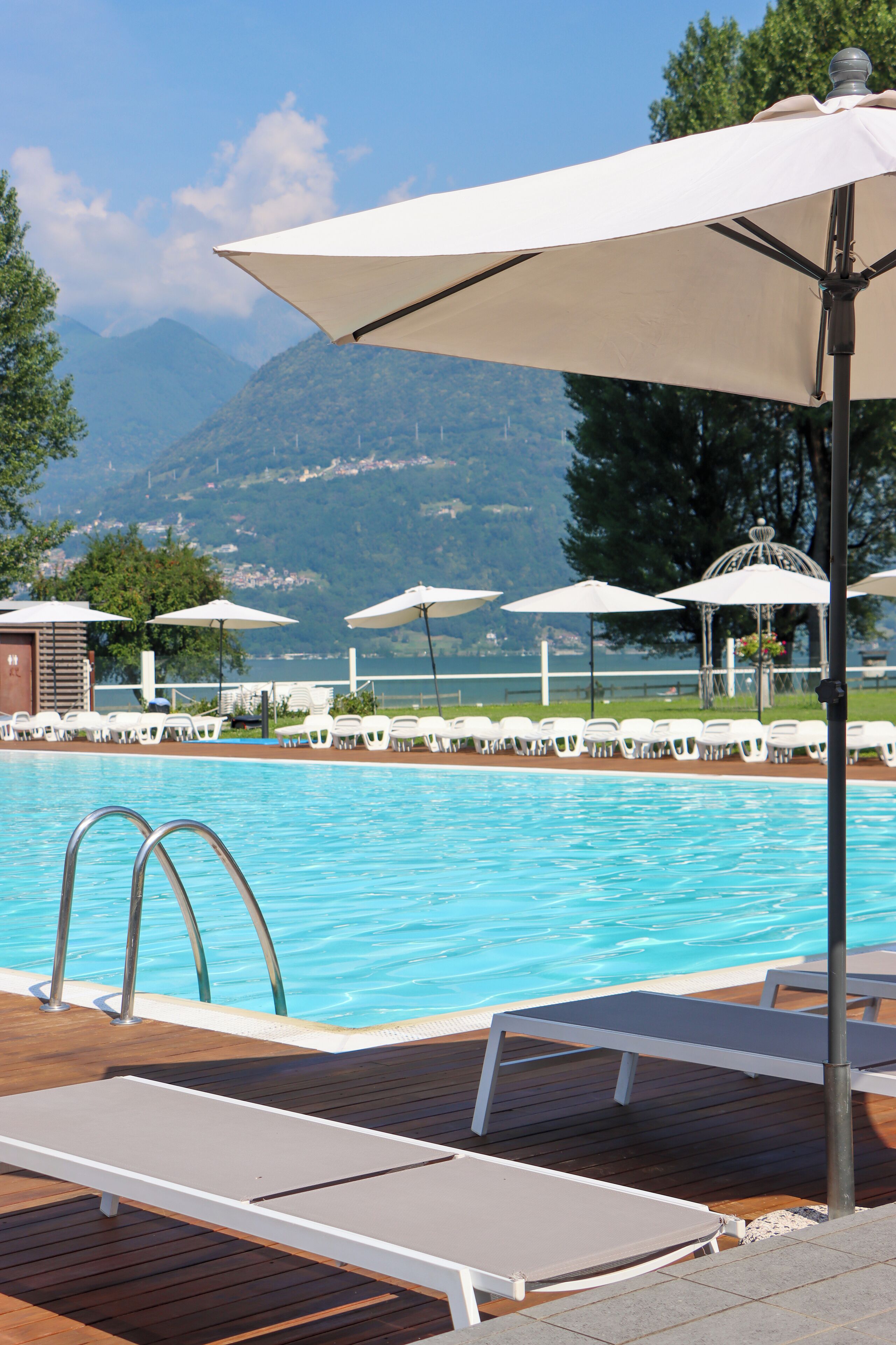 Ein Poolbereich mit Liegestühlen und Sonnenschirmen, eine Holzterrasse und ein Blick auf die Berge im Hintergrund.
