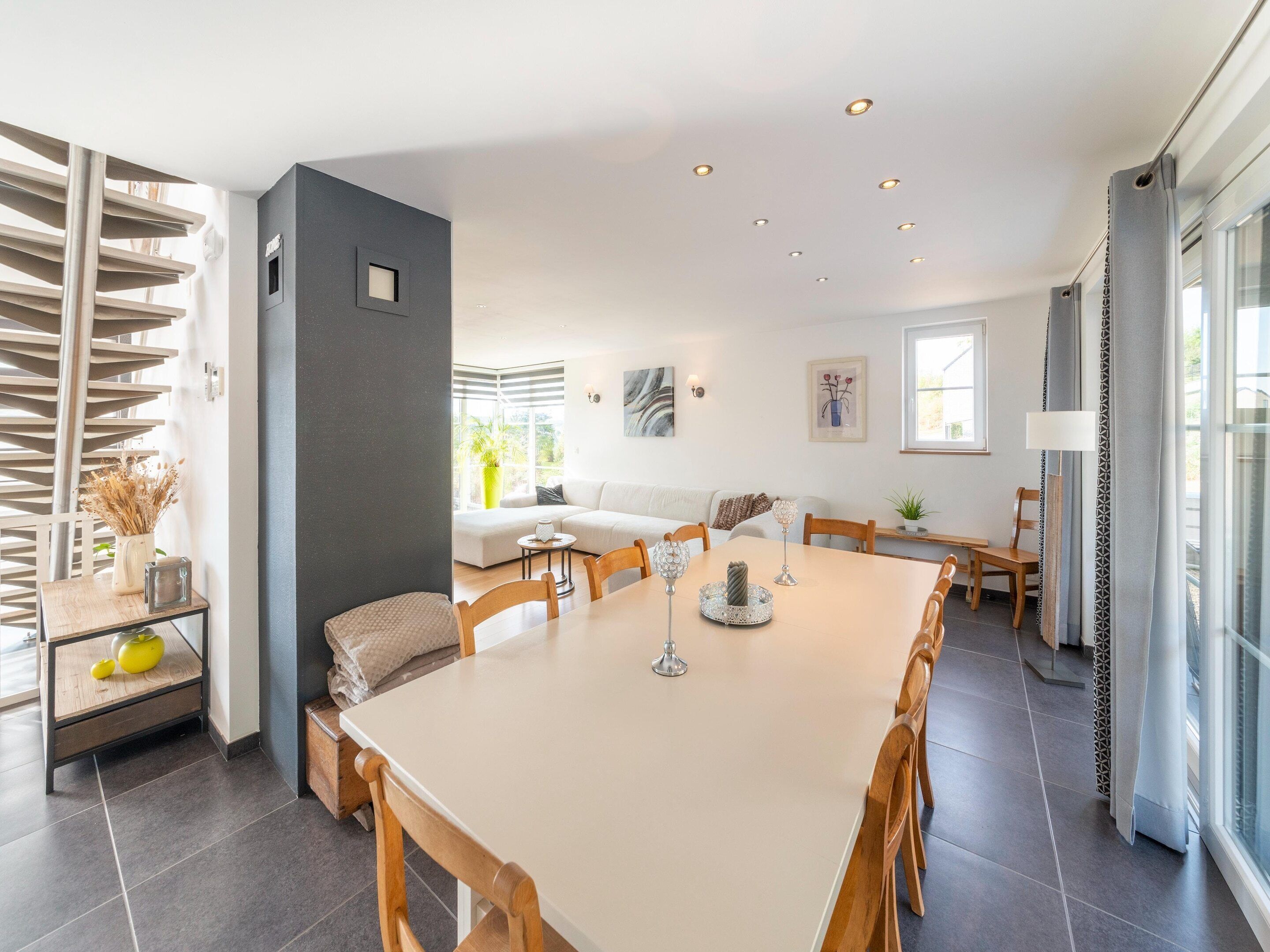 Een moderne eetruimte met een grote tafel, stoelen en een keuken op de achtergrond.