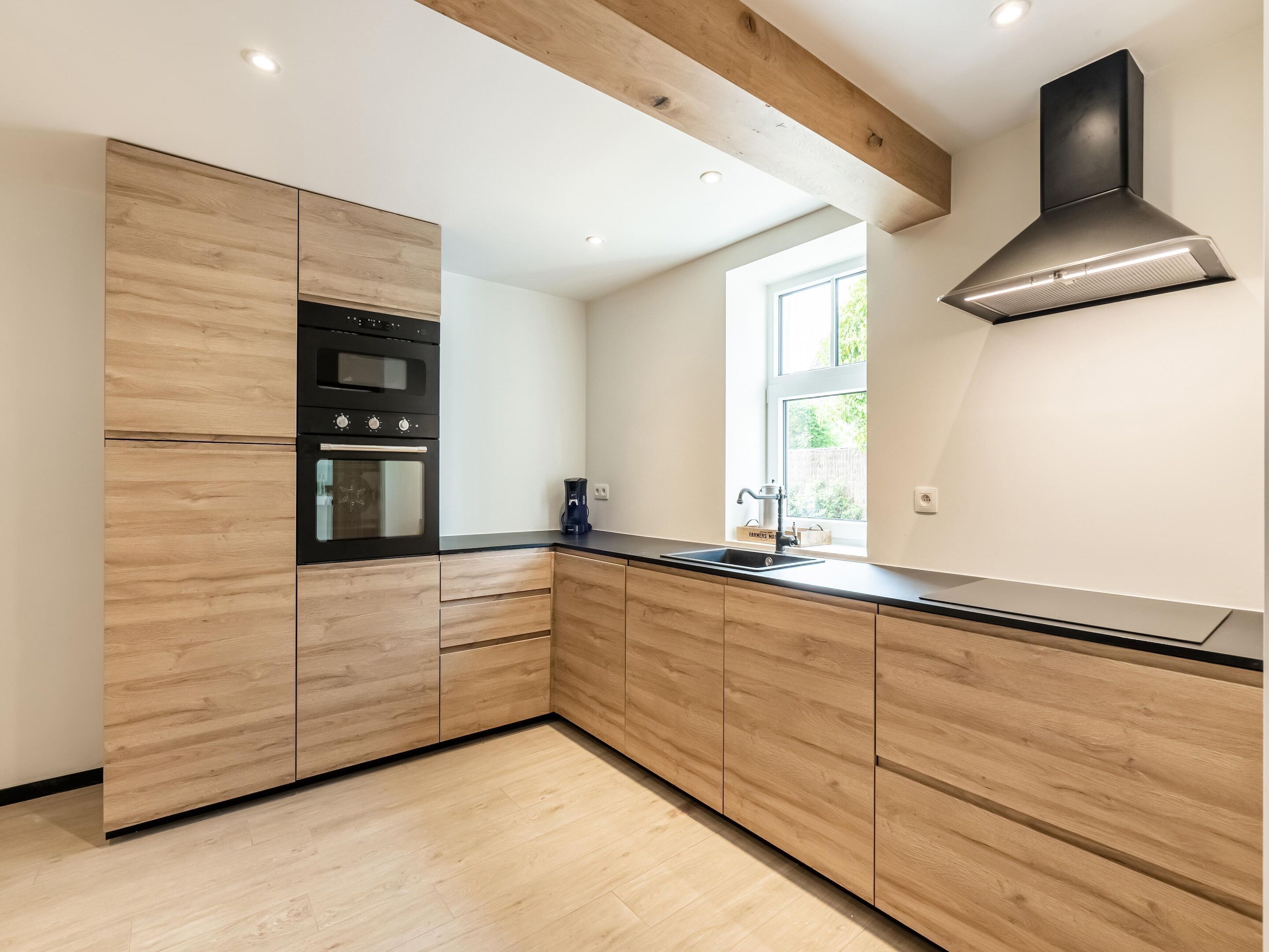 A modern kitchen with wooden cabinetry, a built-in oven, a coffee maker, and a window providing natural light.