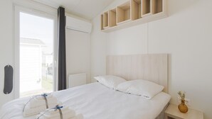 A hotel room with a large bed, a wooden headboard, a bedside table with a vase, and a window with curtains. - Middelkerke Mobile Home With Garden (Middelkerke)