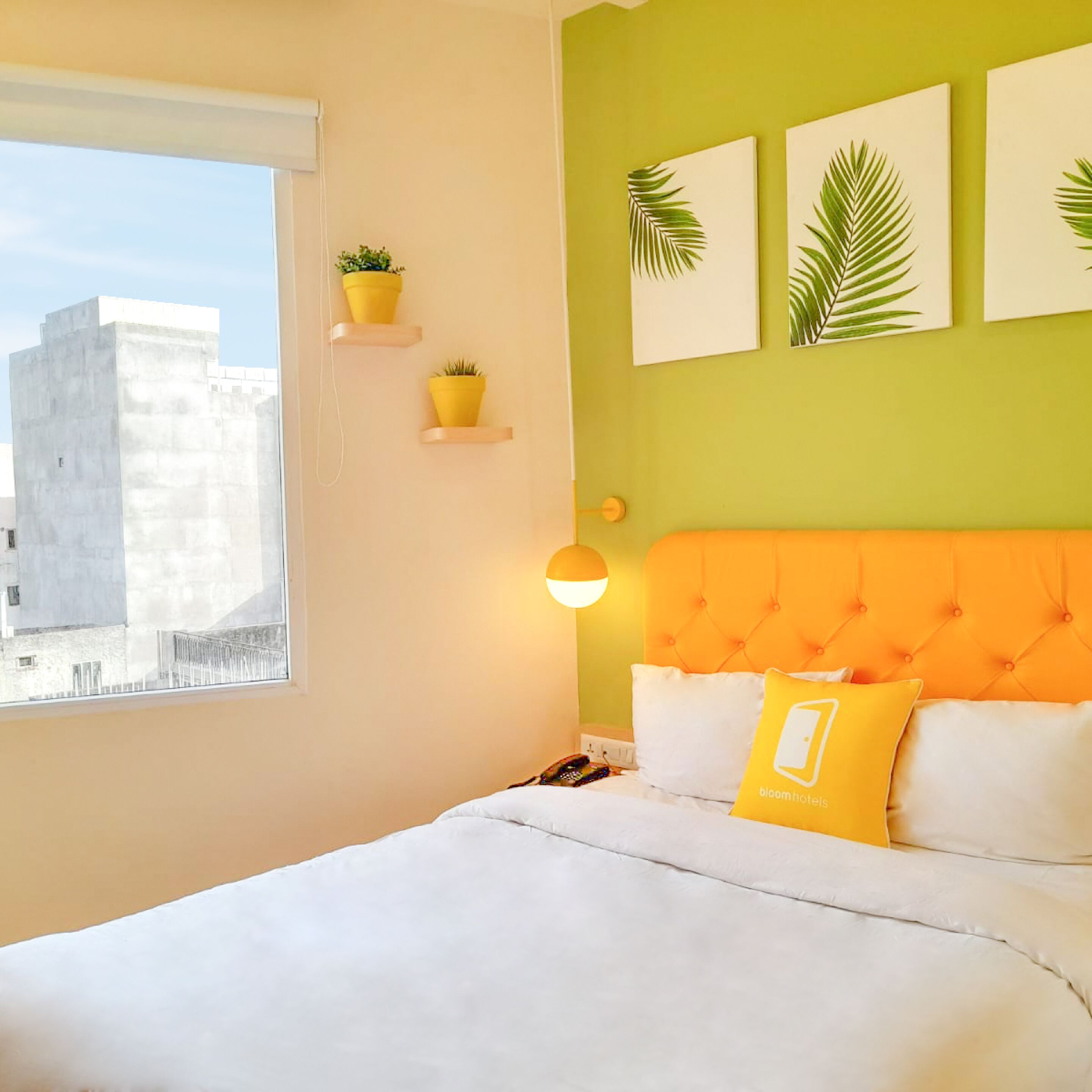 A hotel room with a bed, a window, a view of buildings, and three potted plants on the wall.