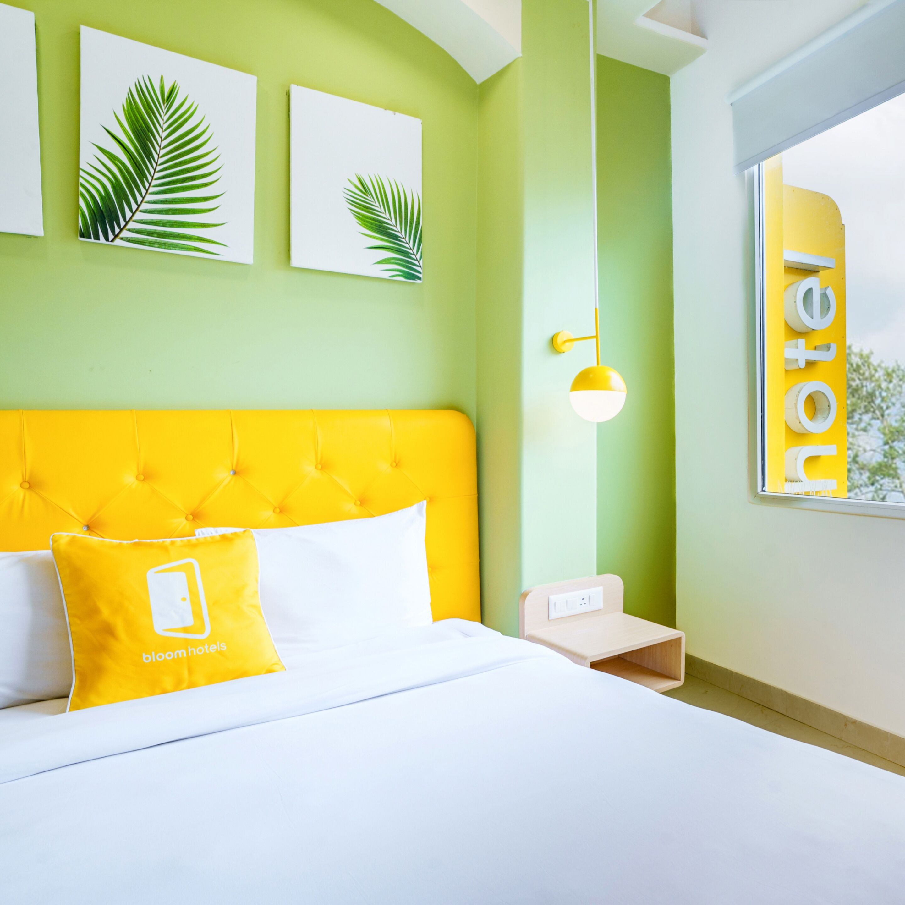 A hotel room with a yellow headboard, white bedding, and a bed with a yellow pillow. There are two framed leaves on the wall, a bedside table with a lamp, and a window with a view of trees.