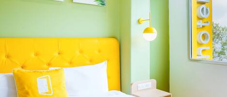 A hotel room with a yellow headboard, white bedding, and a bed with a yellow pillow. There are two framed leaves on the wall, a bedside table with a lamp, and a window with a view of trees.