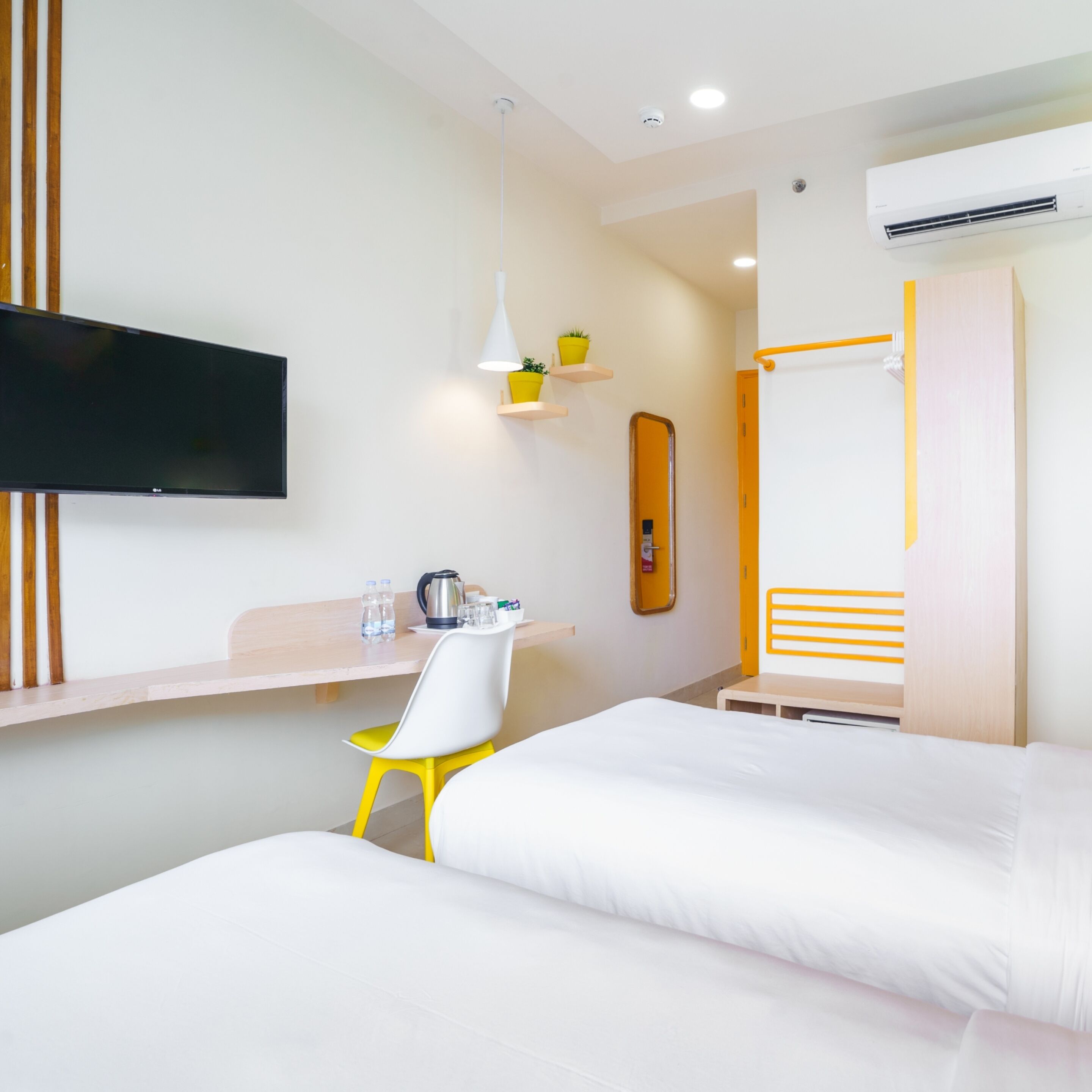 Ein Hotelzimmer mit zwei Betten, einem Schreibtisch mit Stuhl, einem an der Wand befestigten Flachbildfernseher, einem kleinen Tisch mit Wasserkocher und Tassen sowie einer Topfpflanze auf einem Regal.