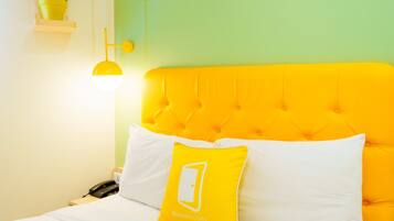 Una cama con cabecera amarilla y ropa de cama blanca, un almohadón amarillo con un logotipo y una lámpara de noche.
