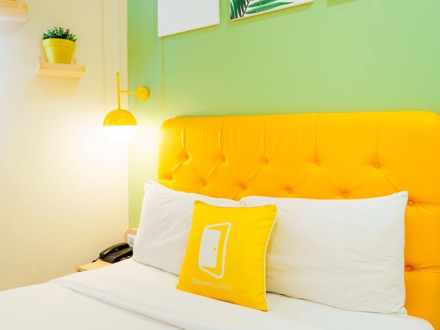 Una cama con cabecera amarilla y ropa de cama blanca, un almohadón amarillo con un logotipo y una lámpara de noche.