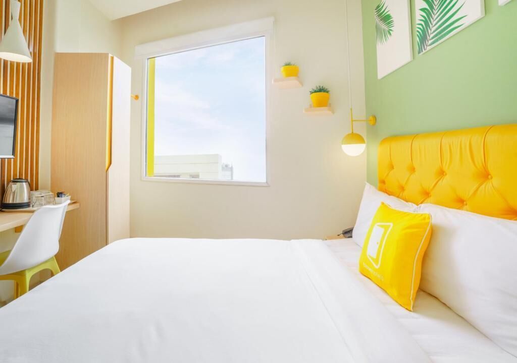 A hotel room with a bed, a desk, a chair, a window, and wall-mounted plants.