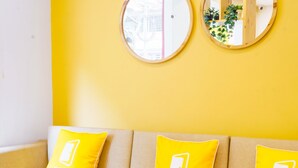 A yellow sofa with three pillows, each featuring a logo. A small plant is on a wooden table in front of the sofa. - Bloom Hotel - Heritage Walk (Amritsar)