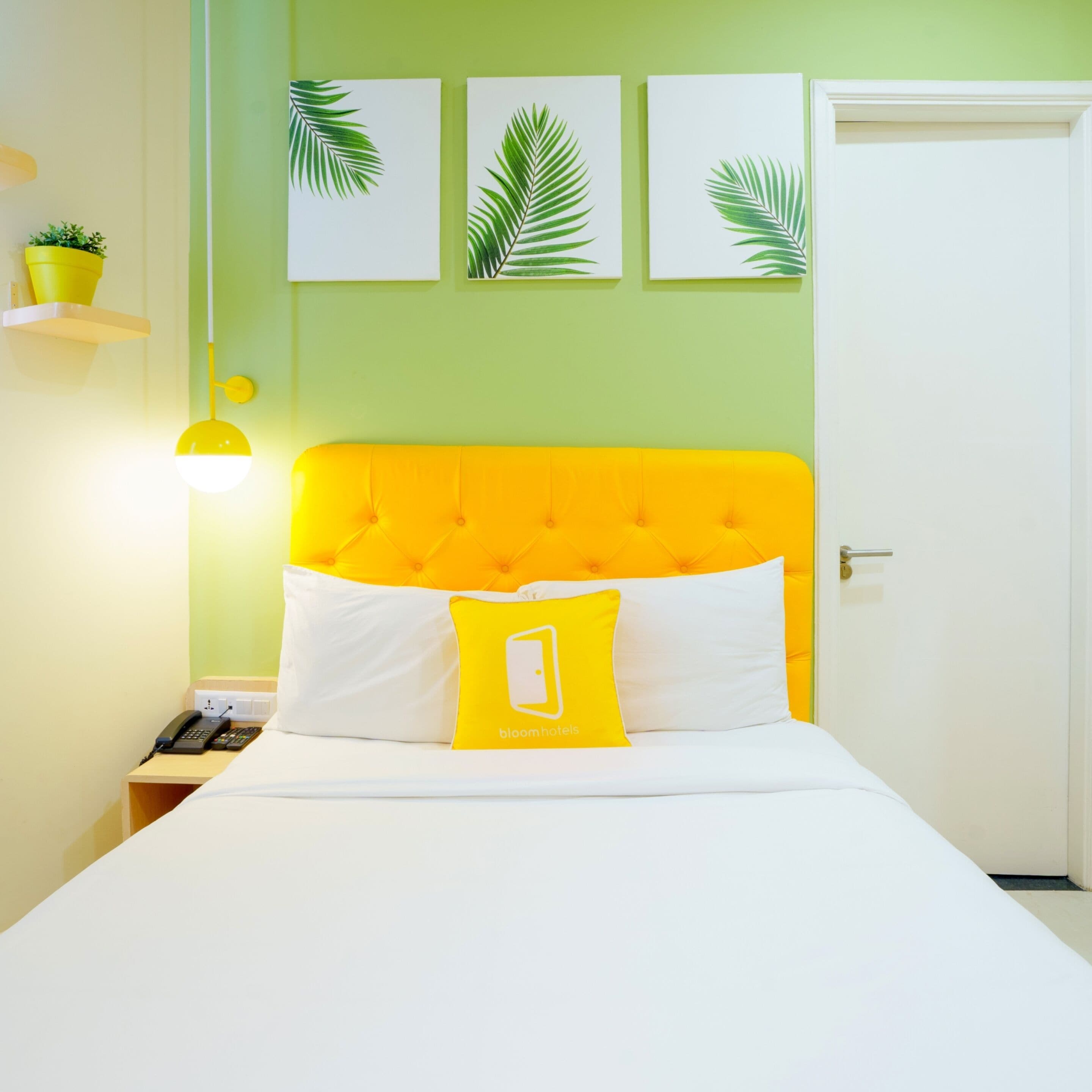 A bed with a yellow headboard, white bedding, and a yellow pillow with a logo. There are three framed leaves on the wall above the bed.