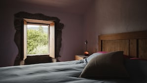 A bedroom with a wooden headboard, a bed with a grey sheet, a nightstand with a lamp, and a window with a view of greenery. - One of two private and intimate casitas. Surrounded by nature. (Moraña)