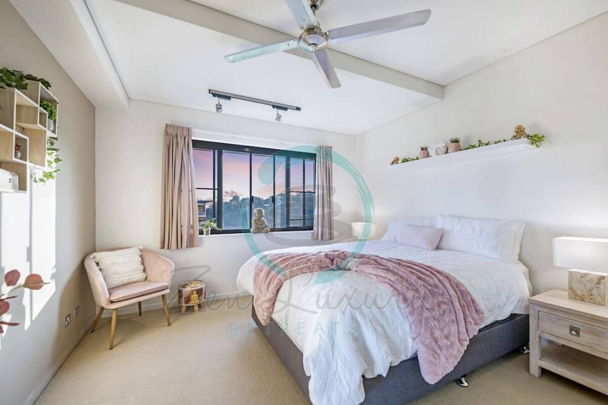 Un dormitorio con cama, una silla, mesita de noche, ventilador de techo, ventana con cortinas y estante con plantas.