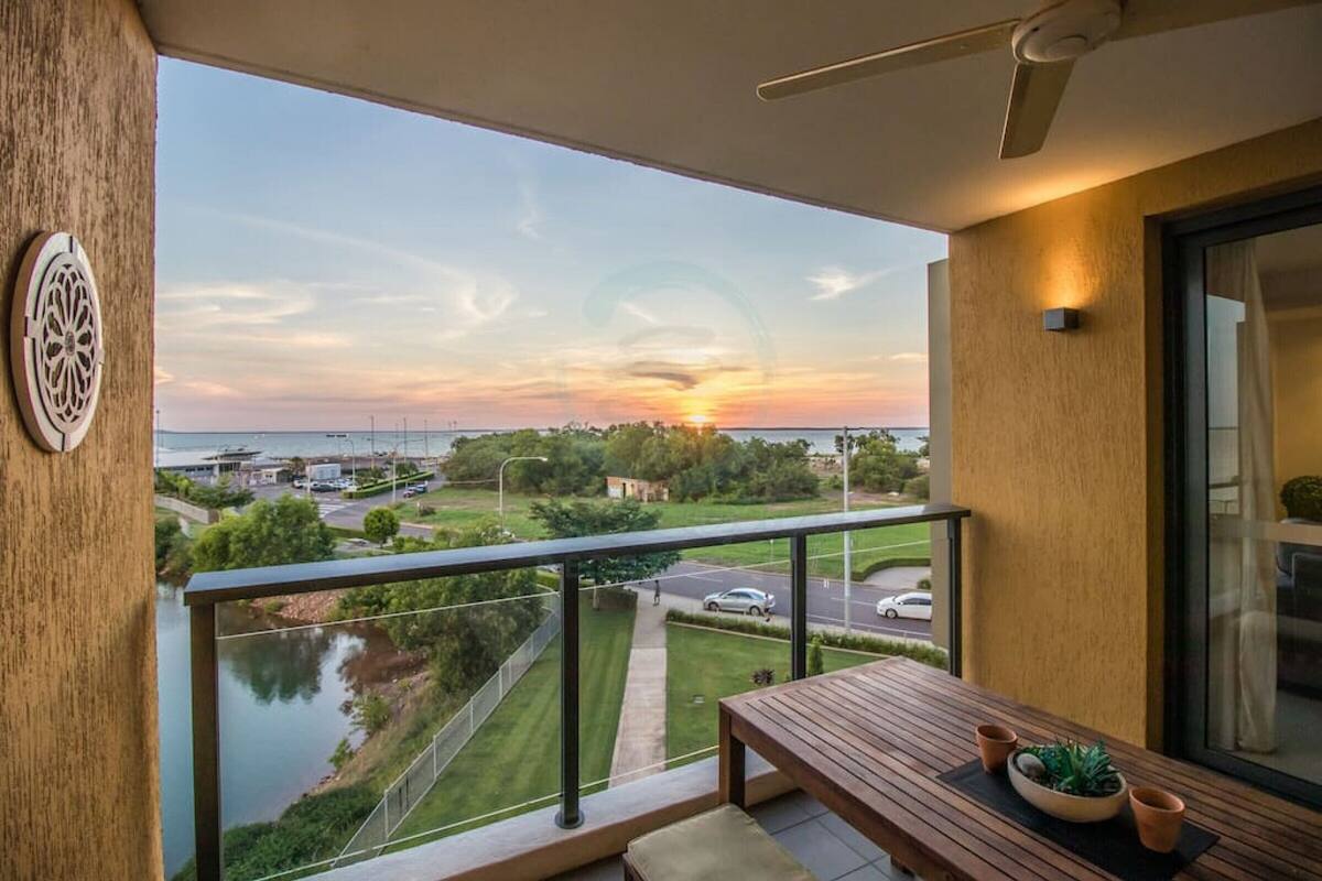 Un balcón con vista a una puesta de sol sobre el agua, una mesa de madera y una planta en maceta.