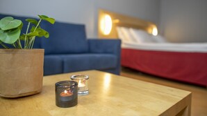 A room with a wooden coffee table, a potted plant, two lit candles, a blue sofa, and a bed with a red bedspread. - Hotelli Uusikuu (Mikkeli)