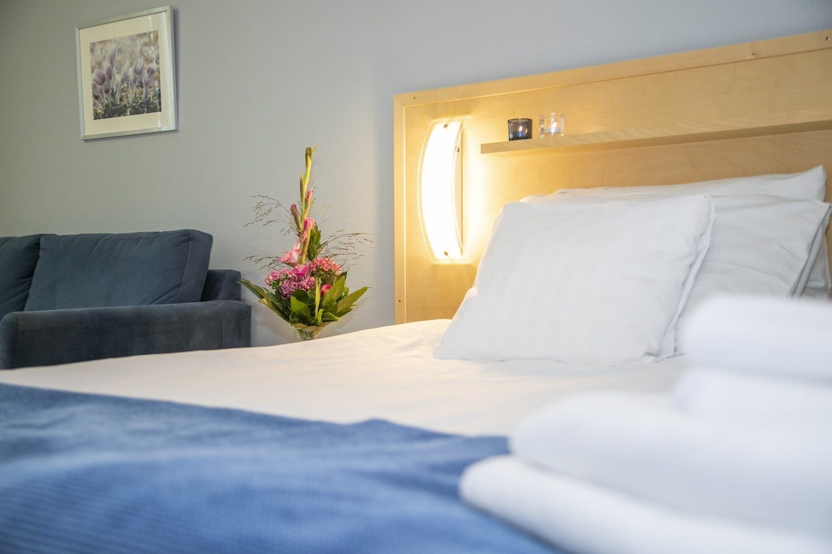 Ein Hotelzimmer mit einem Bett, einem Nachttisch mit Lampe, einem kleinen Tisch mit Blumenvase und einer Couch.