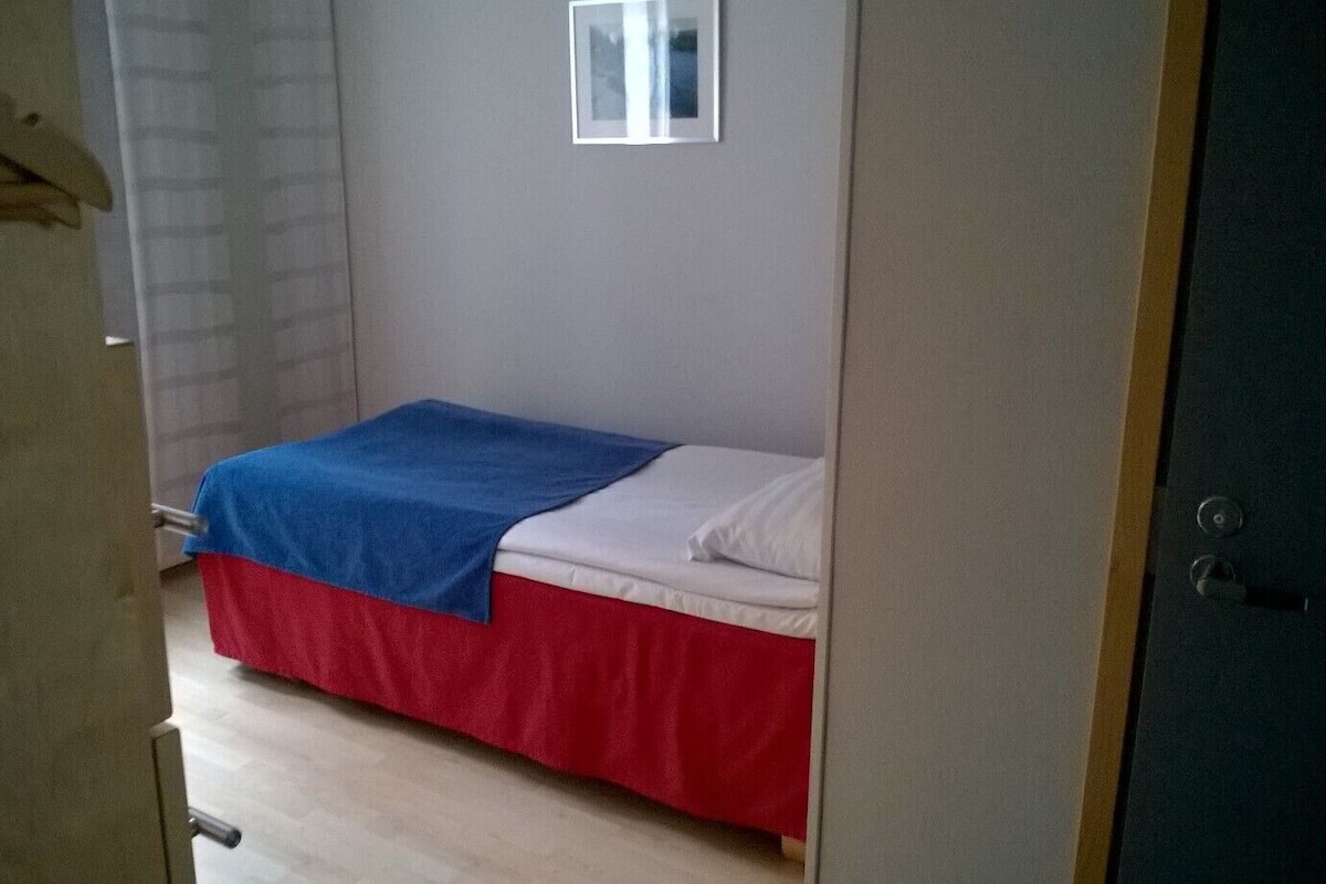 Ein kleines Zimmer mit einem Einzelbett, einer blauen und roten Bettdecke, einem weißen Kissen und einer schlichten Wand mit einem kleinen Bilderrahmen.