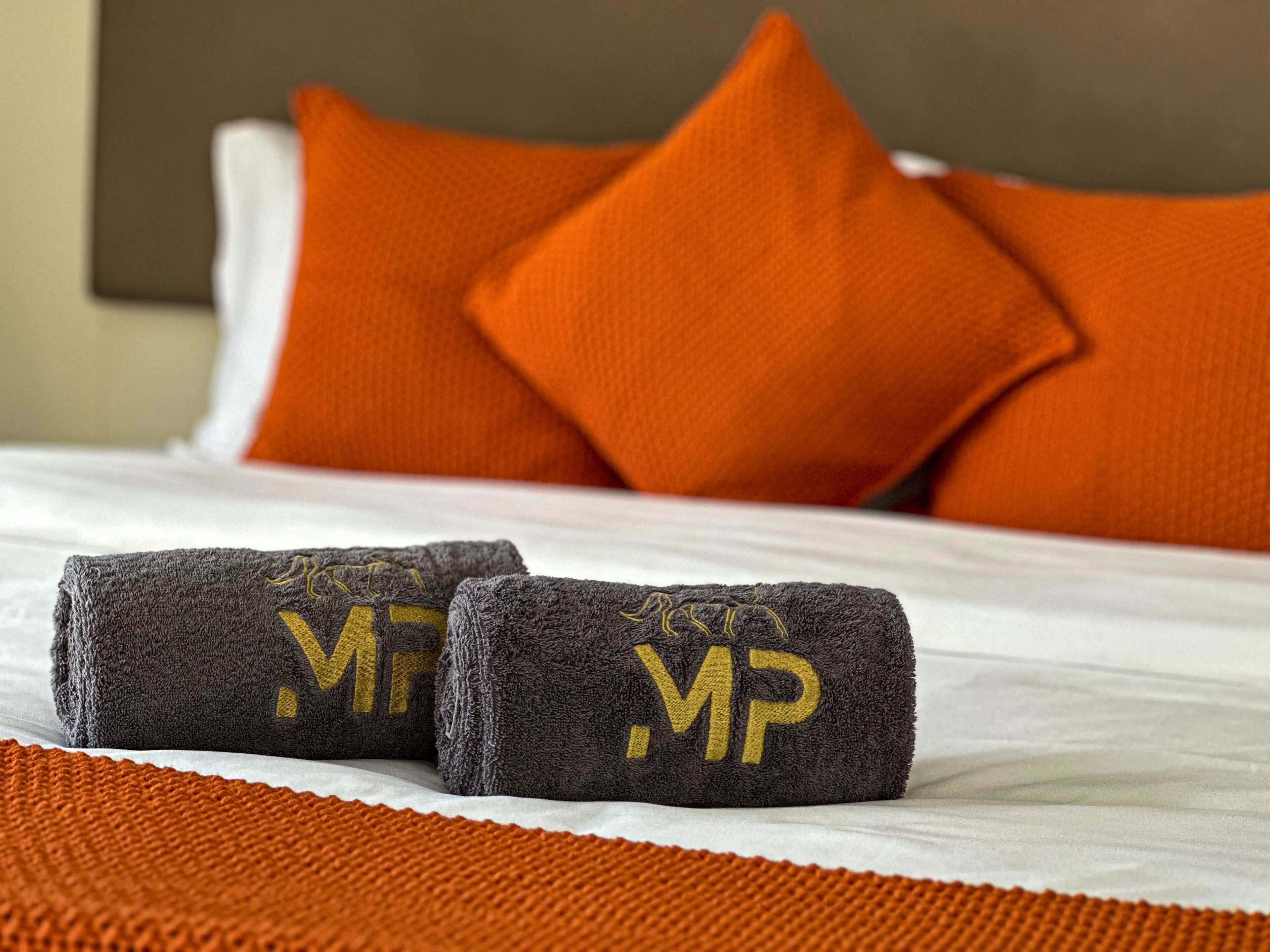 Ein ordentlich bezogenes Bett mit zwei zusammengerollten Handtüchern und zwei orangefarbenen Kissen.