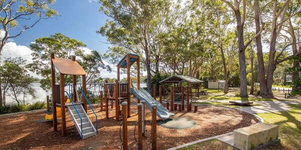 Explore a shaded playground surrounded by mature trees, featuring a slide, climbing frames, and a shelter.