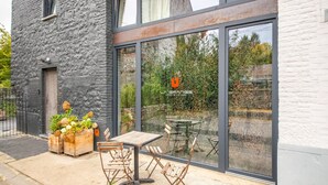A modern building with a stone facade and large glass windows. Inside, there is a wooden table and chairs set up on a concrete patio. - Charming Holiday Home Near Namur With Private Sauna (Jemeppe-sur-Sambre)
