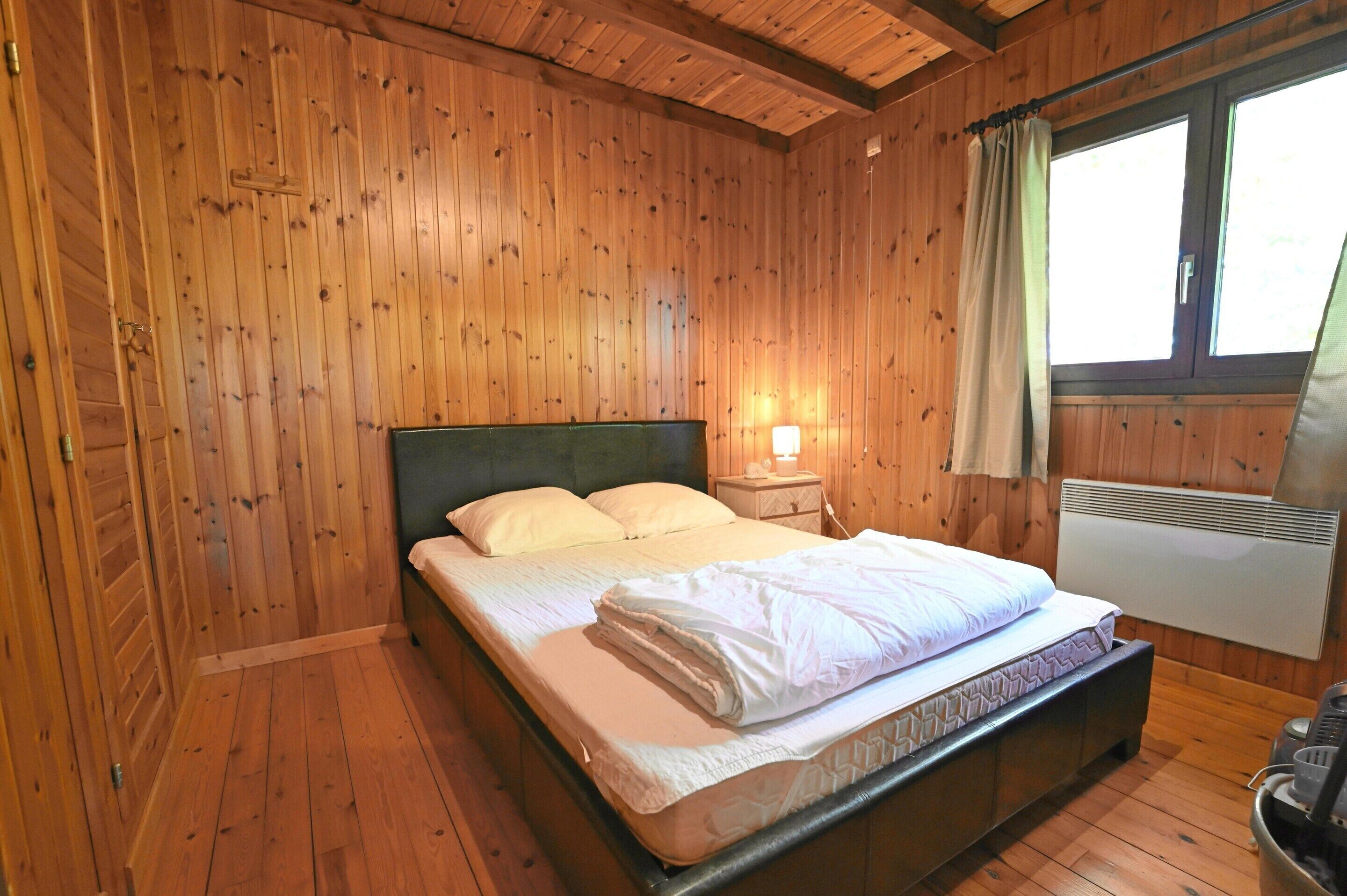 A wooden-paneled room with a bed, a nightstand, a lamp, and a window with curtains.