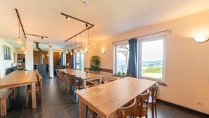 Un vaste espace dédié à la salle à manger, avec une longue table en bois et des chaises, un éclairage suspendu au plafond et une grande fenêtre offrant une vue sur l’extérieur.
