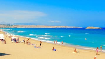A beach with people sunbathing and swimming, clear blue water, and distant mountains.