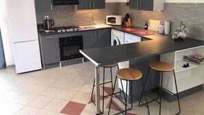 A modern kitchen with grey cabinets, a central island with stools, a built-in oven, a microwave, and a washing machine. - Les Vieilles Ombres - Pineau (Bagnizeau)
