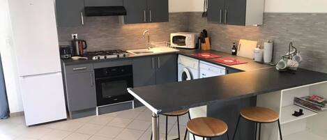 A modern kitchen with grey cabinets, a central island with stools, a built-in oven, a microwave, and a washing machine.