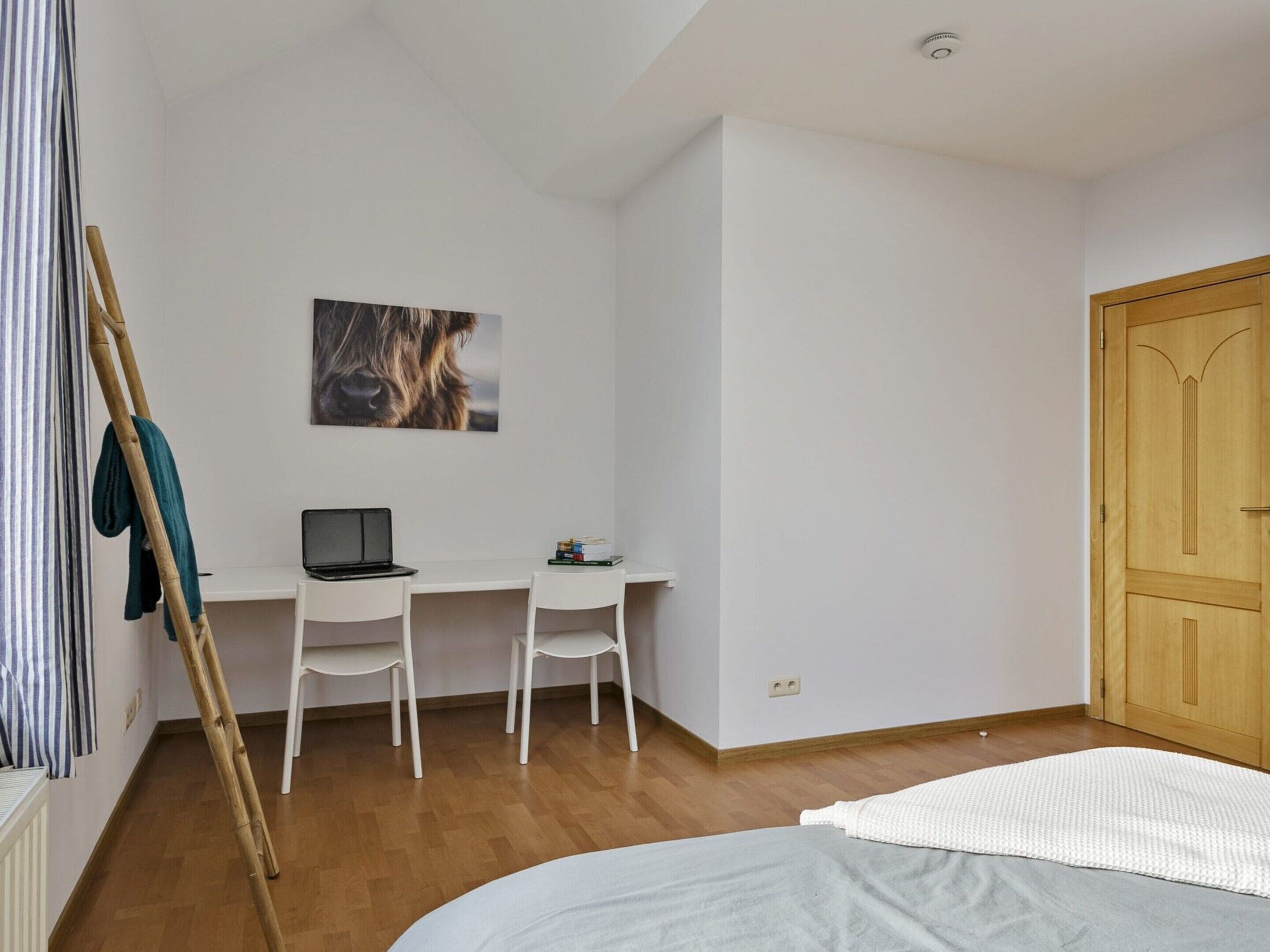 A bedroom with a bed, a desk with a laptop, two chairs, a wooden wardrobe, and a framed picture on the wall.