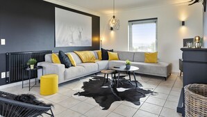 A modern living room with a grey sofa, yellow cushions, a black and white cowhide rug, a black chair, and a black and gold pendant light. - Pleasant Holiday Home With Recreation Room (Bouillon)
