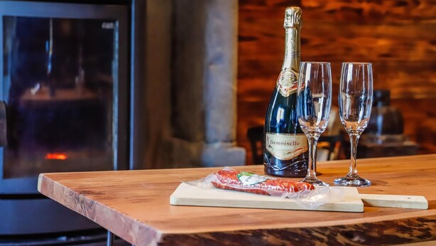 A wooden table with a bottle of wine and two glasses, a wooden cutting board with a wrapped item, and a fireplace in the background. - Quietly Located Farmhouse L'echappee Belle (Manhay)