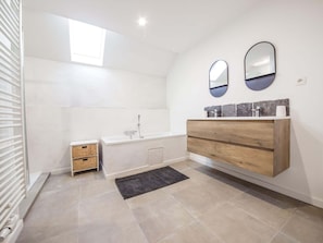 Ein modernes Badezimmer mit einer freistehenden Badewanne, einem hölzernen Waschtisch mit Marmorplatte, zwei Wandspiegeln und einem kleinen Korbwannenstuhl.